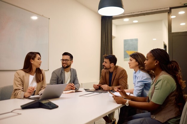 A young team of five people conversing around a table at work, showing how a disc profile helps build a good team.