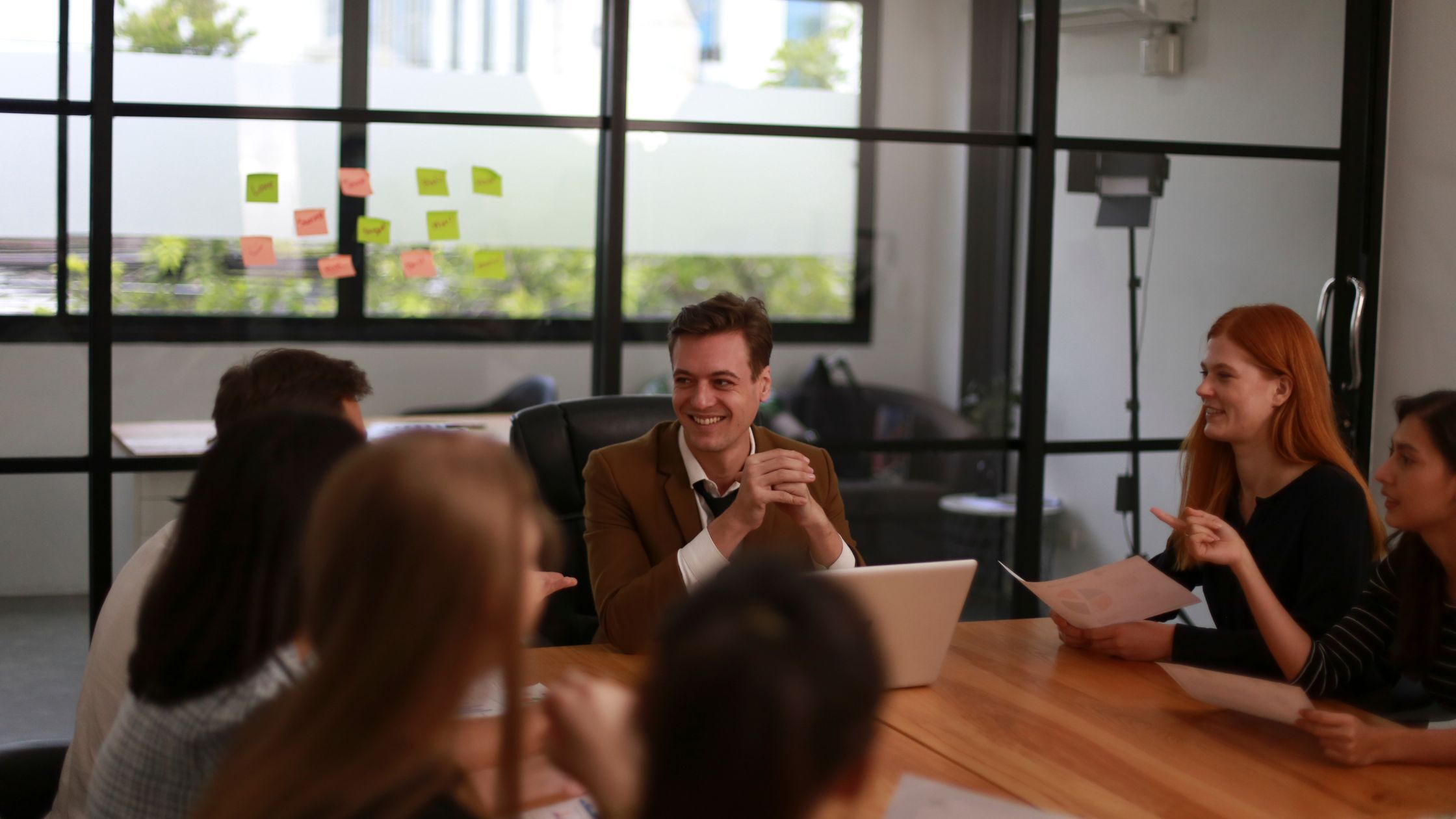 A diverse professional team in a bright UK office setting engaged in a collaborative meeting around a wooden table, discussing printed reports.